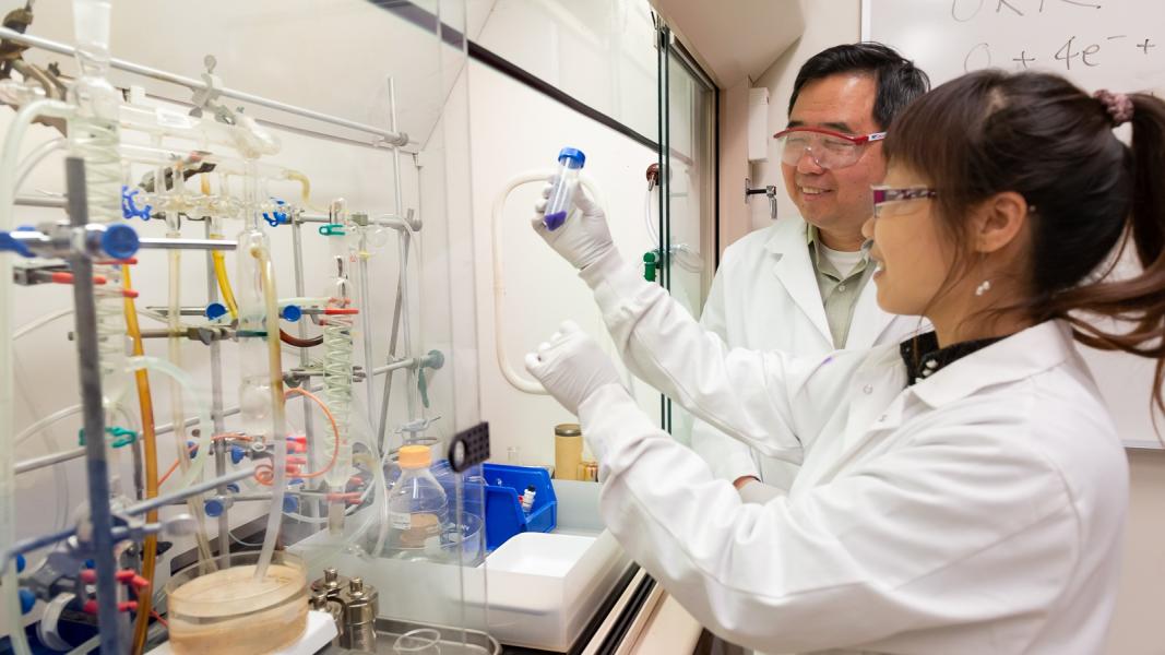 Two goggled scientist in white lab coats and gloves. One is holding capped glass tube of dark substance.