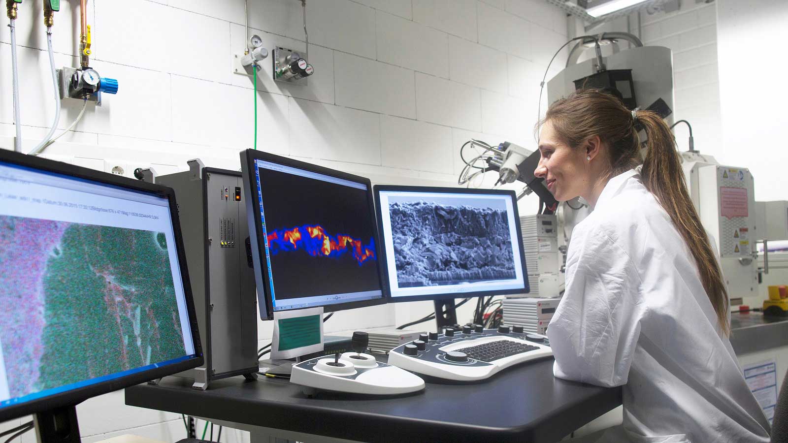 Woman in lab coat closely scrutinizing two monitors while seated at desk.