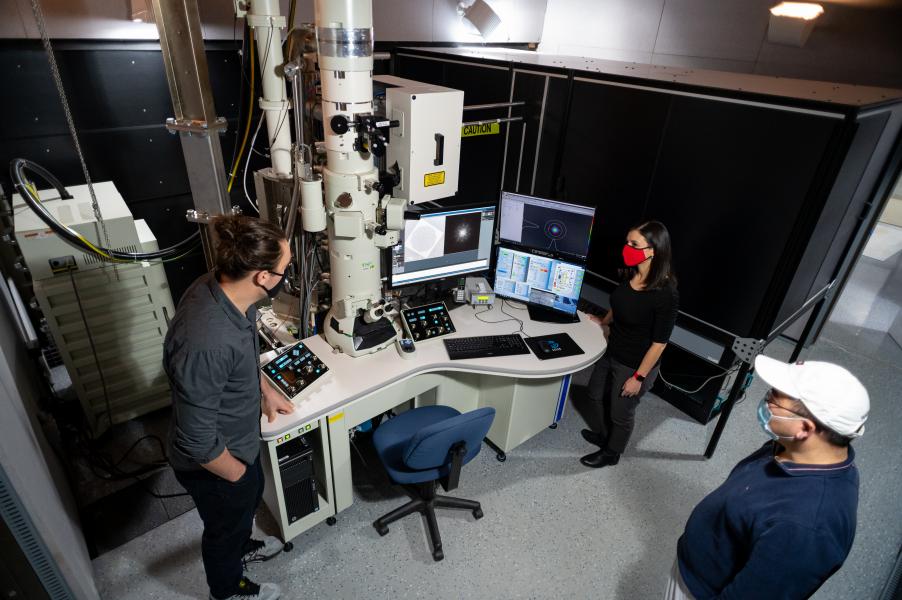 Three people with face masks standing around desk with tall microscope and computer monitors.